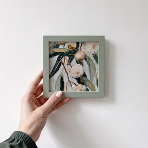 May include: A small, square painting in a sage green frame. The artwork depicts a close-up of eucalyptus branches with green leaves and white, fluffy flowers. The painting is held by a person.