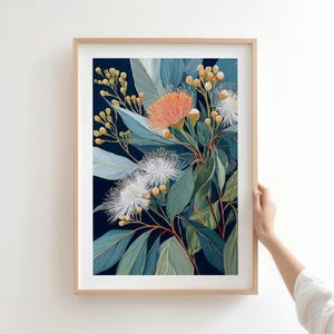 Puede incluir: Una impresi&oacute;n bot&aacute;nica enmarcada con un arreglo floral. La obra de arte muestra una mezcla de tonos verdes, azules y naranjas, con representaciones detalladas de hojas, capullos y delicadas flores blancas y naranjas. El marco es de color madera clara.
