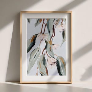 May include: A framed art print depicting eucalyptus branches with long, pale green leaves and delicate, light pink flowers. The artwork is set against a soft, light blue background and framed in a natural wood frame.