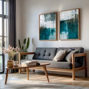 Może przedstawiać: Nowoczesny salon z szarą sofą futon z drewnianą ramą, uzupełniony drewnianym stolikiem kawowym i dwoma abstrakcyjnymi dziełami sztuki w drewnianych ramach. Pokój jest udekorowany roślinami i miękkim oświetleniem.