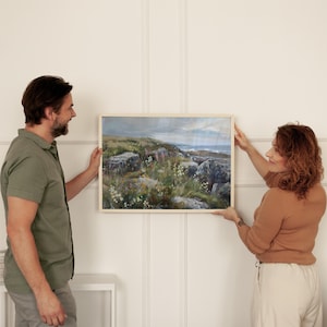 Könnte beinhalten: Ein Paar hängt ein gerahmtes Landschaftsbild an einer weißen Wand. Das Bild zeigt eine felsige Küste mit grünem Gras und Wildblumen.