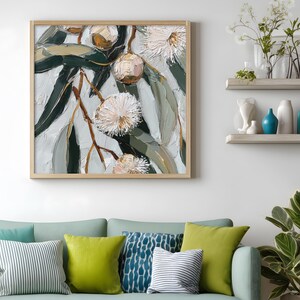 May include: Square framed artwork featuring a close-up of eucalyptus blossoms and leaves. The painting uses a palette of white, green, and brown, with textured brushstrokes. The artwork is displayed above a light blue sofa with decorative pillows.