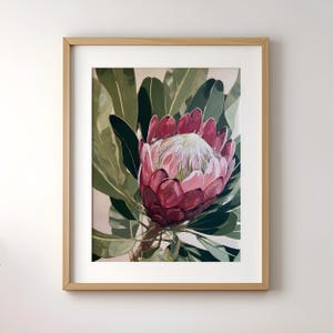 Può includere: Stampa floreale incorniciata con un grande fiore di protea. Il fiore ha un centro rosa intenso con petali bianchi e rosa chiaro. Grandi foglie verdi circondano il fiore, su uno sfondo rosa chiaro. La cornice è color legno chiaro.