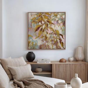Op de afbeelding: Een ingelijst abstract schilderij met een botanisch thema. Het kunstwerk toont olijfgroene en bordeauxrode bladeren, met witte en roze accenten. Het schilderij hangt boven een houten kast met decoratieve vazen en boeken.