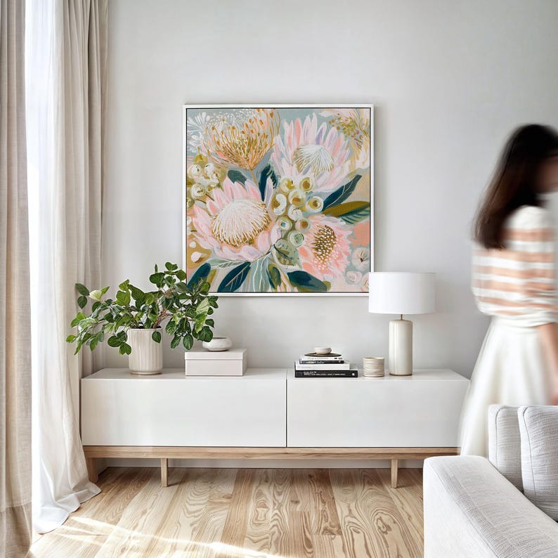 May include: A square framed floral painting with a white border. The artwork features large pink and peach flowers with green leaves and accents. The painting hangs above a white cabinet with a wooden base, a lamp, and a plant.