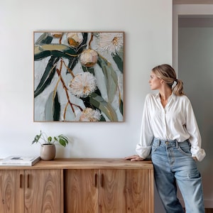 May include: Square framed floral painting featuring white, yellow, and green flowers and leaves. The artwork is displayed on a light-colored wall above a wooden cabinet. The painting has a textured, impasto style.