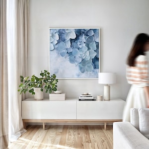 Pode incluir: Um console de mídia branco com duas gavetas e uma lâmpada branca em um piso de madeira. Uma pintura abstrata azul está pendurada na parede acima do console. Uma planta fica no console.