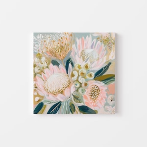 May include: Square canvas art featuring a floral arrangement. The artwork showcases large protea flowers in shades of pink, peach, and gold, with green and blue-green leaves. The background is a soft, muted blue-green.