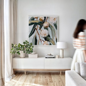 May include: A framed floral painting with white and green leaves and flowers hangs above a white cabinet. A white lamp and a potted plant are on the cabinet. The room has a neutral color palette with a wooden floor.