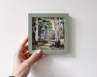 May include: A framed landscape painting featuring a forest scene with tall trees and a path. The artwork is in a square, sage green frame. The painting depicts a serene outdoor scene with a variety of trees and foliage.