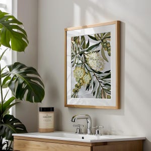 May include: A framed wall art print featuring a botanical illustration of cream-colored banksia flowers and green foliage. The artwork is displayed in a light wood frame with a white mat, hanging above a bathroom sink with a chrome faucet.
