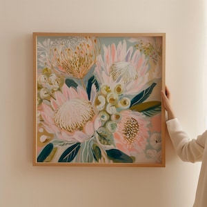 May include: A framed floral art print featuring a bouquet of protea flowers in shades of pink, peach, and gold, with green and blue-green leaves. The artwork is set against a light blue background and framed in a natural wood.