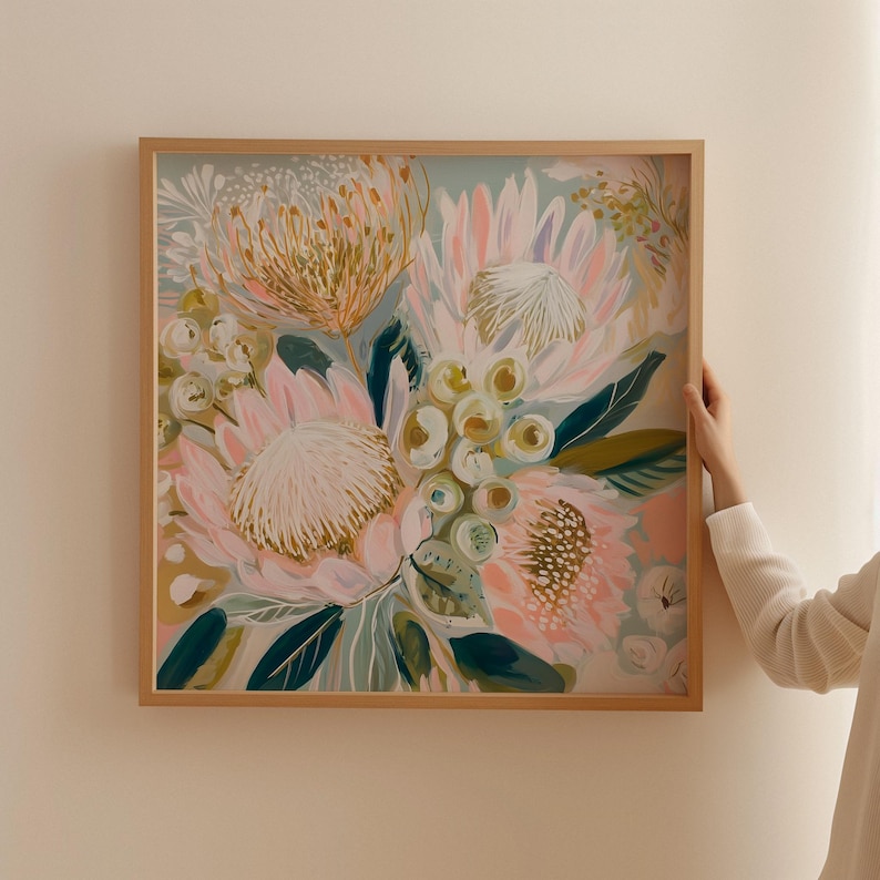 May include: A framed floral art print featuring a bouquet of protea flowers in shades of pink, peach, and gold, with green and blue-green leaves. The artwork is set against a light blue background and framed in a natural wood.