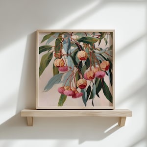 Op de afbeelding: Een ingelijste botanische print met eucalyptus takken met groene bladeren en roze en gele bloemen. Het kunstwerk wordt tentoongesteld op een licht houten plank tegen een witte muur. De lijst is van licht hout.