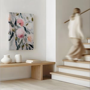 以下が含まれることがあります： ピンクと白の花と緑の葉が描かれたキャンバスプリント。2つの白い花瓶と2冊の本が明るい木製のベンチに置かれています。背景には、階段を降りるぼやけた人物がいます。