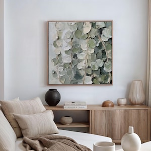 May include: Square framed artwork featuring a textured painting of cascading eucalyptus leaves in shades of green and white. The artwork hangs above a wooden cabinet with decorative vases and books, creating a serene, natural aesthetic.