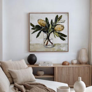 May include: A framed painting featuring yellow flowers in a clear glass vase, with green leaves, set against a neutral backdrop. The artwork hangs above a wooden cabinet, adorned with various decorative objects.