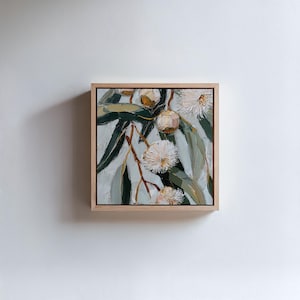 May include: Square framed oil painting of eucalyptus branches with green leaves, white flowers, and brown seed pods. The artwork is set against a light gray background and framed in natural wood.