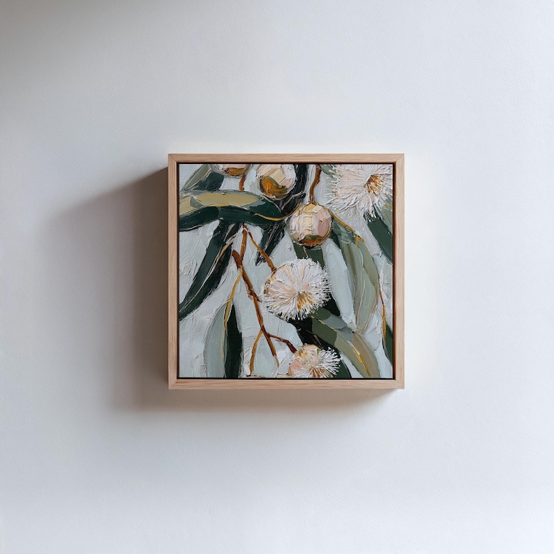 May include: Square framed oil painting of eucalyptus branches with green leaves, white flowers, and brown seed pods. The artwork is set against a light gray background and framed in natural wood.