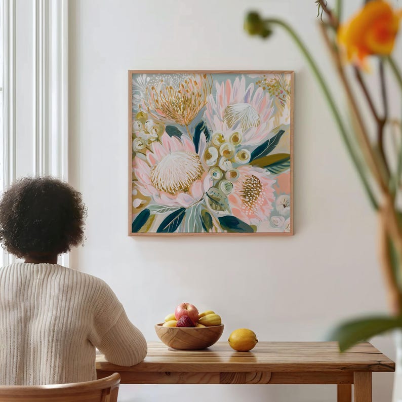 May include: A framed floral painting featuring large, pink and cream-colored protea flowers with green and blue foliage. The artwork is displayed on a white wall above a wooden table. A wooden bowl of fruit sits on the table.