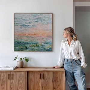 Puede incluir: Pintura abstracta de paisaje en un marco de madera. La obra presenta una mezcla de colores, incluyendo azules, verdes y naranjas, creando un efecto texturizado. La pintura se exhibe sobre un mueble de madera con una planta en maceta.