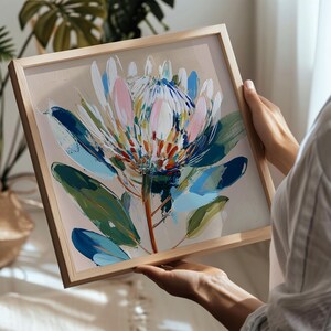 Könnte beinhalten: Gerahmter Kunstdruck mit einer farbenfrohen Protea-Blume. Das Kunstwerk zeigt eine lebendige Mischung aus Blau, Grün, Rosa und Weiß vor einem neutralen Hintergrund. Der Holzrahmen verleiht dem Stück eine natürliche Note.