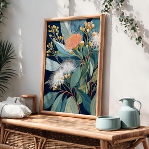 Puede incluir: Una impresi&oacute;n bot&aacute;nica enmarcada que presenta un vibrante arreglo floral con flores naranjas, blancas y amarillas, sobre un fondo azul oscuro. La obra de arte se exhibe en un banco de madera junto con una jarra y un cuenco azul claro, creando una escena serena inspirada en la naturaleza.