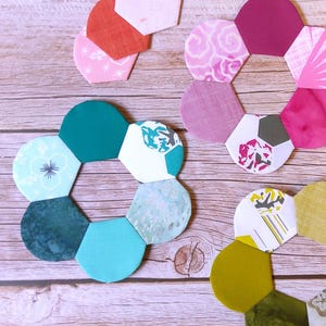 May include: A collection of fabric hexagons arranged into three circular flower-like shapes and several loose pieces. The fabric pieces feature various colors including teal, magenta, olive green, orange, and white, with assorted floral and geometric patterns.