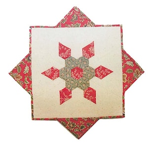 May include: A handmade quilt block featuring a central star design. The star is composed of red and green floral patterned fabric, set against a cream-colored background. The block is framed by a red floral patterned border.