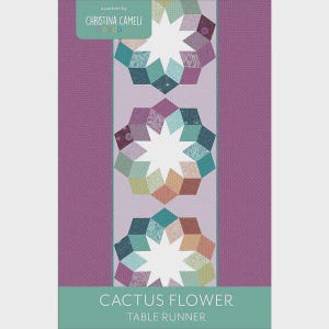 May include: A quilt pattern titled Cactus Flower Table Runner by Christina Cameli. The design features three star-shaped motifs in shades of teal, purple, orange, and green, set against a light purple center panel with dark purple side borders.
