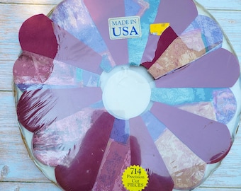 May include: A circular jigsaw puzzle in shrink wrap featuring a flower-like arrangement of petal-shaped pieces in shades of purple, maroon, and blue. A white label reads MADE IN USA and a yellow star-shaped sticker reads 714 Precision Cut PIECES.