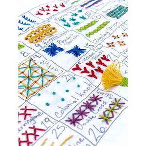 May include: A white fabric sampler featuring a grid of various hand-embroidered stitches in multiple colors. Each square contains a specific stitch pattern labeled with handwritten text, including names like Colonial knot, Lattice, and Russian chain stitch.