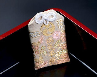 Puede incluir: Un amuleto omamori de tela beige con bordados florales en dorado, rosa y blanco. La parte superior está adornada con un cordón blanco anudado. El centro muestra un emblema circular dorado sobre caracteres kanji japoneses verticales.