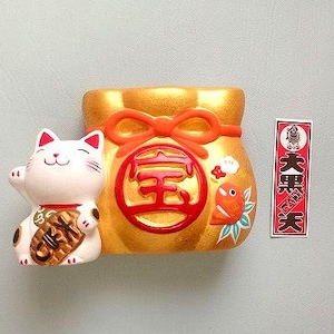 以下が含まれることがあります： 片手を上げた招き猫の陶器製置物。赤いリボンと日本の文字が付いた金色の巾着袋の隣にあります。日本語のテキストが入った赤い長方形の看板も付いています。