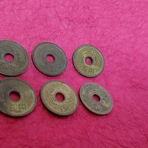 以下が含まれることがあります： 中央に穴と浮き彫りの文字がある、6枚の日本のヴィンテージ5円硬貨。硬貨はブロンズ色で、質感のあるマゼンタ色の表面に配置されています。これらの硬貨は収集品です。