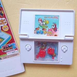 May include: White Nintendo DS Lite handheld game console with a colorful screen featuring Mario, Yoshi, and other characters. The game console is open, revealing a water game. A game box is visible on the left.