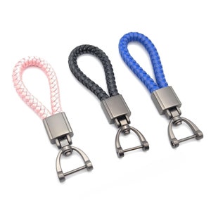 Puede incluir: Tres llaveros trenzados con diseño de lazo en rosa claro, negro y azul. Cada llavero incluye un conector rectangular de metal oscuro y un cierre giratorio en forma de D con una barra horizontal para asegurar las llaves de forma firme.