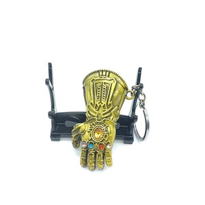 May include: A gold-colored metal keychain shaped like Thanos's gauntlet with infinity stones. The gauntlet is attached to a black metal keychain ring.