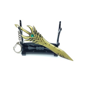 Puede incluir: Un llavero de espada de color dorado con una gema verde en la empuñadura, exhibido en un soporte negro.