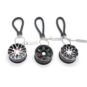 Könnte beinhalten: Drei Schlüsselanhänger mit Miniatur-Auto-Felgen, schwarzen geflochtenen Lederbändern und silbernen Metallringen. Jede Felge hat ein einzigartiges Speichendesign, darunter ein komplett schwarzes Sechs-Speichen-Modell und zwei silber-schwarze Vielspeichen-Modelle. Jeder Anhänger enthält ein kleines rechteckiges Metallschild mit der Gravur hellaflush.