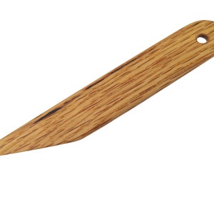 Puede incluir: Una herramienta de madera con una punta puntiaguda y un mango redondeado con un agujero. La herramienta está hecha de madera marrón claro con vetas marrón oscuro.