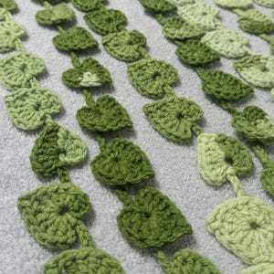 May include: A close-up of a crocheted garland featuring multiple strands of green heart-shaped leaves. The leaves are crocheted in varying shades of green, creating a gradient effect.