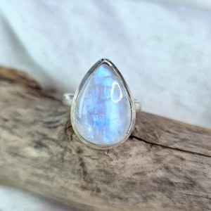 Puede incluir: Anillo de plata con una piedra lunar iridiscente en forma de pera. La piedra tiene un brillo azul y blanco, engastada en un bisel de plata. El anillo se muestra sobre madera flotante sobre un fondo de tela blanca.