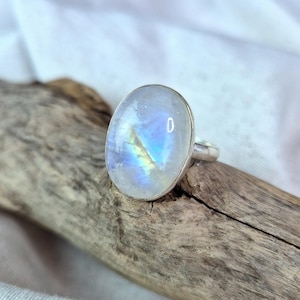 Puede incluir: Anillo de plata con una gran piedra lunar ovalada. La piedra lunar tiene una base blanca lechosa con destellos iridiscentes azules y amarillos. El anillo está colocado sobre un trozo de madera flotante sobre un fondo de tela blanca.