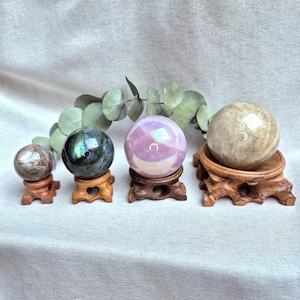 Könnte beinhalten: Vier Kristallkugeln in verschiedenen Farben und Größen auf Holzständern. Die Kugeln sind aus Labradorit, Amethyst, Selenit und Calcit.