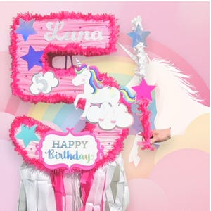 May include: Pink unicorn-themed piñata shaped like the number 5. The piñata is decorated with pink and white fluffy trim, a unicorn, stars, and the name "Luna". The piñata also has a sign that says "Happy Birthday".