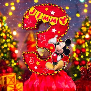 May include: A red and yellow cardboard number three with a Mickey Mouse character and the name "Jimmy" on a banner. The number is decorated with gold stars and is set against a blurred background of Christmas lights and trees.