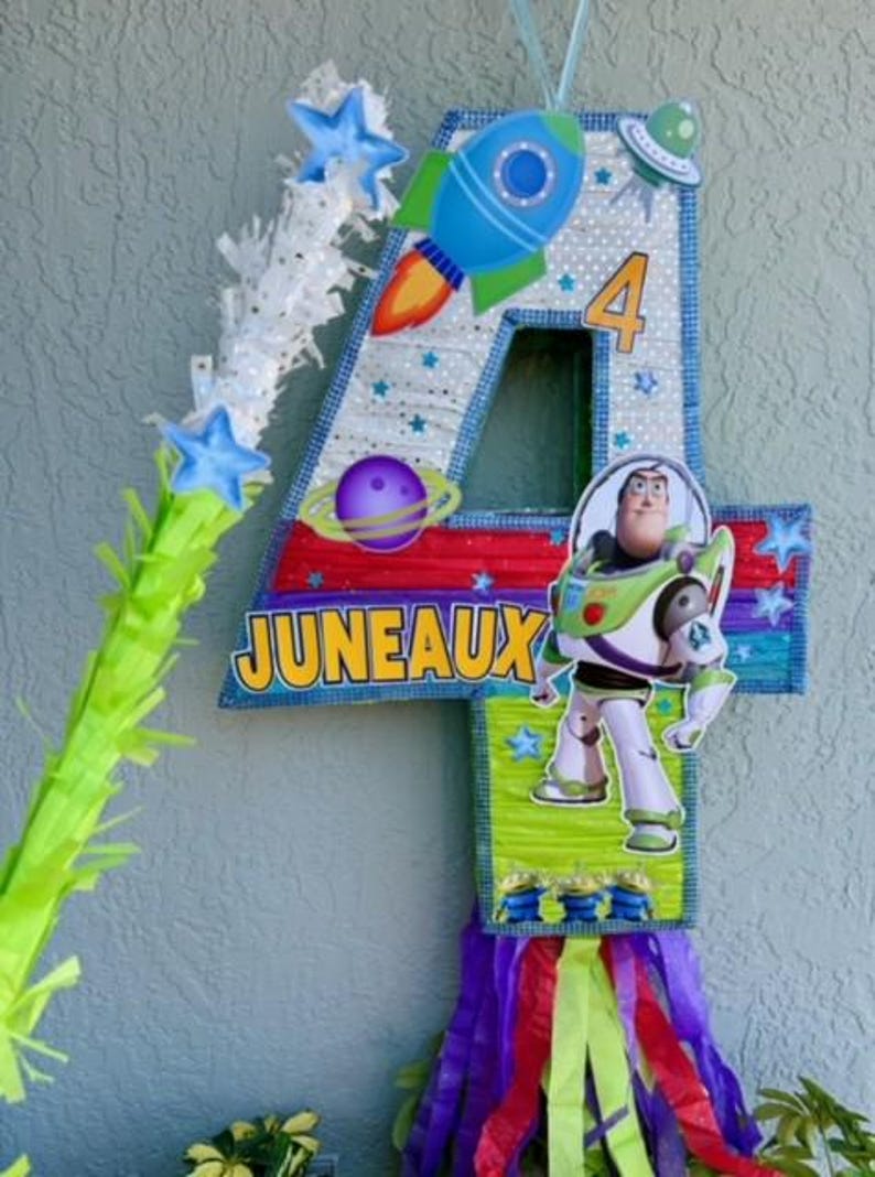 Puede incluir: Una colorida pi&ntilde;ata hecha a mano con forma de n&uacute;mero 4, decorada con un tema espacial. Presenta un cohete, estrellas, planetas y un personaje de la pel&iacute;cula Toy Story. La pi&ntilde;ata est&aacute; etiquetada como "JUNEAUX" y tiene serpentinas de colores que cuelgan de la parte inferior.