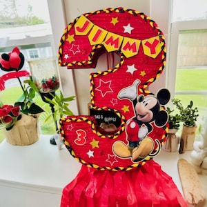May include: A red and yellow Mickey Mouse themed pi&ntilde;ata shaped like the number three. The pi&ntilde;ata has a banner that says "Jimmy" and features Mickey Mouse in a red and yellow outfit.