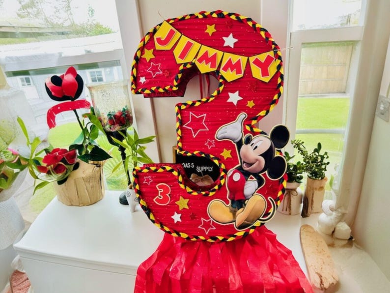 May include: A red and yellow Mickey Mouse themed pi&ntilde;ata shaped like the number three. The pi&ntilde;ata has a banner that says "Jimmy" and features Mickey Mouse in a red and yellow outfit.